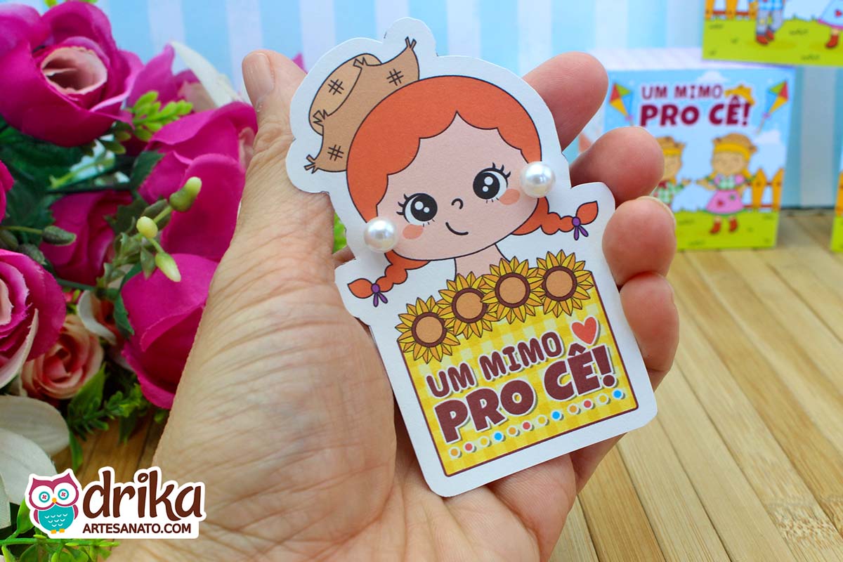 Cartão de menina caipira ruiva com brincos de pérola, na mão, e flores rosas ao fundo.
