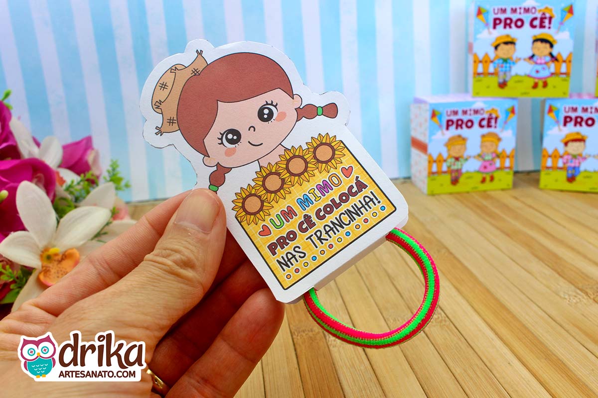 Porta xuxinha de cabelo menina caipira com cabelo castanho em mãos, com texto "Um mimo pro cê colocá nas trancinha" e xuxinha verde e rosa.